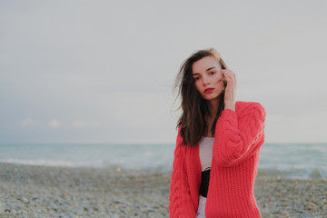 Sad brunette girl in red cardigan alone on empty seashore in windy weather