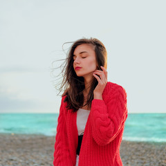 Sad brunette girl in red cardigan alone on empty seashore in windy weather. Wind ruffles hair