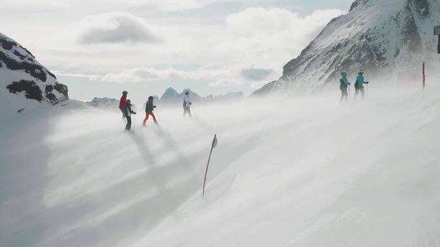 Aerial shot of group of skiers on top of snowy mountain range as wind blows snow over top between them.