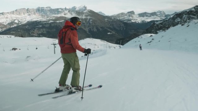 Following male tourist skier downhill in Italy ski resort. Mountain range view in background.