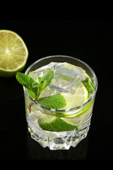 Glass of cocktail with rum, lime, ice cubes and mint leaves on black mirror background.