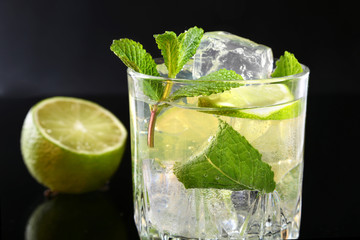 Glass of cocktail with rum, lime, ice cubes and mint leaves on black mirror background.