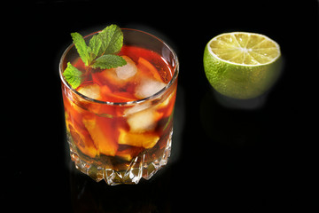 Glass of Dark Rum Cocktail with lime, orange, ice cubes and mint leaves on black mirror background.