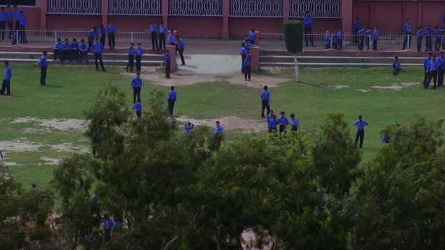 School students playing in the ground aerial view passing the trees, students are in blue pants and shirts,  some part is missing of grass in the ground, school building in the background