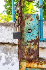 Heavy rust on a painted lock of a metal door missing a door handle with a rusty chain and an open padlock