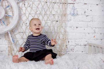 Fototapeta premium Portrait of a cute and naughty european little girl in a sailors clothing in the studio of marine style on a background of white brick wall