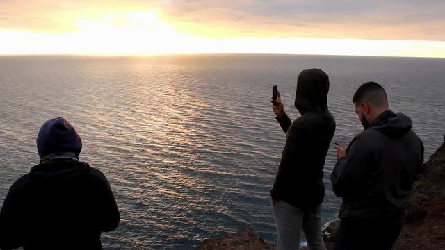 Three men standing on cliff edge enjoying sunset by the sea in Gran Canaria, Spain. Group of friends taking photos of splendid twilight with cellphone in Canary Islands. Travel concept