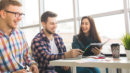 Group of international university students having fun studying in library, three colleagues of modern work co-working space talking and smiling while sitting at the desk table with laptop computer