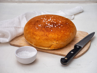 Homemade bread on a wooden unpainted board with coarse salt in a white salt shaker and a kitchen knife, next to it is a white cotton napkin