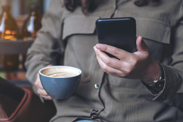 Closeup image of a woman holding , using and looking at smart phone while drinking coffee in cafe