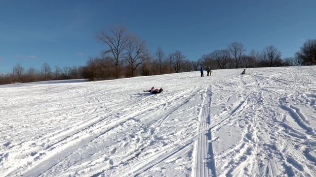 A kid sliding downhill on a plastic board. Vividly excited and having the time of his life, in slow motion. It's obviously over excited to spend time outdoors with his friends and siblings.