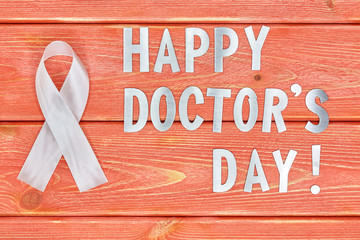 white awareness ribbon and inscription of iron letters: Happy doctors day. lying on wooden textured...