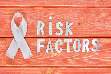 white awareness ribbon and inscription of iron letters: risk factors, lying on wooden textured background color of season 2019