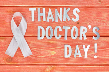 white awareness ribbon and inscription of iron letters: thanks doctors day lying on wooden textured background color of season 2019