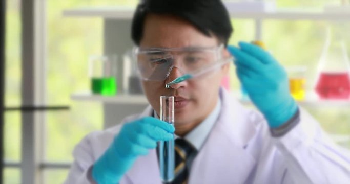 Good looking Asian scientist doing experiment in laboratory, he dropping blue liquid into test tube, colorful test tubes on table behind him.