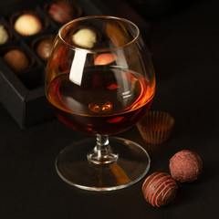 A glass of brandy and chocolates in a paper box. Alcoholic drink on a dark background.Selective focus. Horizontal frame. Dark photo. Square frame