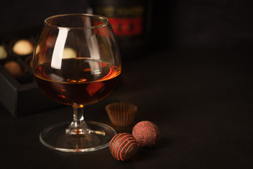 A glass of brandy and chocolates in a paper box. Alcoholic drink on a dark background. Selective focus.