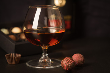 A glass of brandy and chocolates in a paper box. Alcoholic drink on a dark background. Selective focus.