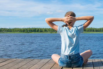 the girl on the background of the sky and water, sitting on the pier, crossed her hands behind her head and sunbathing, on the left there is a place for the inscription on the clouds