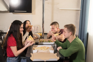 Company of four young friends sitting in kitchen and enjoying eating pizza