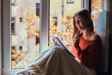 Beautiful girl in a warm sweater covered her legs with a blanket, makes notes in her notebook sitting on the window sill next to the open window
