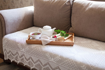 White teapot, mug of mint healthy beverage in cozy home interior. Drinks, book with flower are on wooden serving tray. Breakfast on sofa with lace blanket in morning sunlight. Still life for tea time.