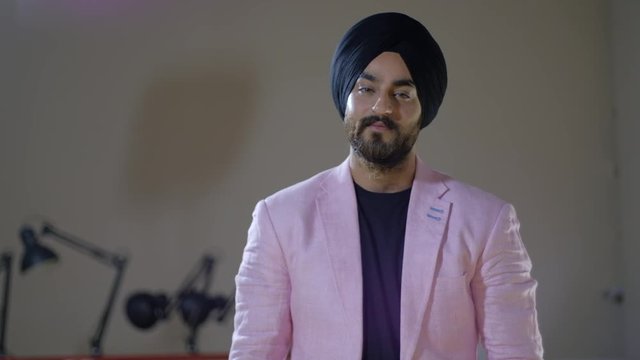 Confident and sharp smart black turbaned man looking at the camera with intent and confidence, smiling smugly and acting smart, in light pink coat jacket