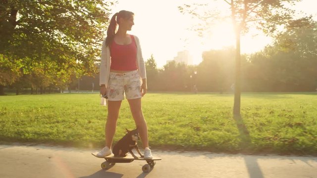 LENS FLARE: Cheerful Caucasian girl and cute puppy riding a high tech electric longboard through the sunlit park. Cool shot of senior dog and its owner skateboarding on a sunny summer afternoon.