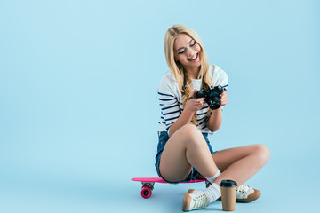 Smiling girl sitting on pink skateboard and holding camera on blue background