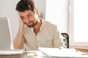 Image of uptight office worker 30s wearing white shirt using laptop and paper documents, while...