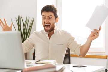 Image of aggressive office worker 30s wearing white shirt, using laptop and paper documents in...