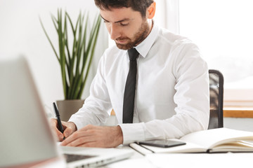 Fototapeta premium Image of successful office worker 30s wearing white shirt working with laptop and notebook, in modern workplace