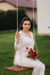 Lady in white sit on swing on the back yard of her house. She hold a re flowers