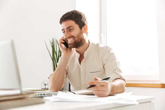 Image of joyful businessman 30s wearing white shirt talking on mobile phone, while sitting at table in modern office