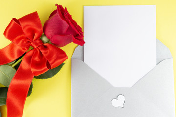 A fresh red rose big bud and petals with green leaves bow near white empty letter in silver open envelope Yellow background Invitation or Felicitation Minimalist concept Copy Space and template