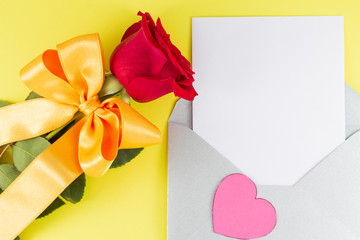 A fresh red rose big bud and petals with green leaves bow near white empty letter in silver open envelope Yellow background Invitation or Felicitation Minimalist concept Copy Space and template