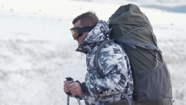 A tourist with trekking poles and a huge backpack against a background of snow-capped mountains in the spring climbs to a top and stops for a halt to drink tea from a thermos. 4K slowmotion. 