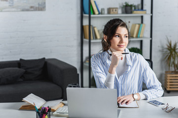 Attractive businesswoman holding pen, using laptop and looking away