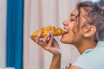 Photo of a cheerful young woman happy. Going to the table and eating croissant.