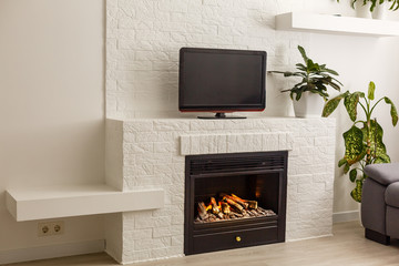 Room in a modern style. There is a TV on the white wall, white rack with a flower in vase, TV remote, decorations in a form black fireplace