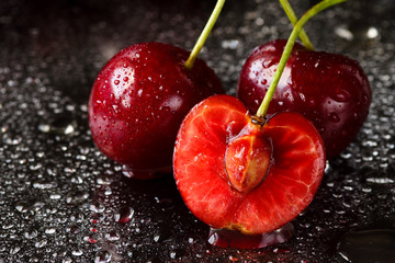 Extreme close up of two and a half sliced fresh, ripe cherry fruits with the stems and pits. Black background with water drops and reflections