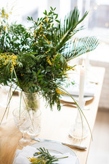Beautiful springtime table setting with green leaves and mimosa branches, bright white table dinner decoration