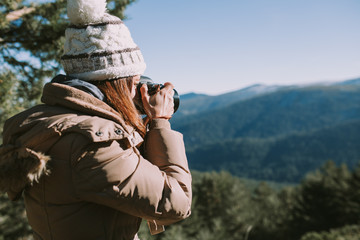 A brunette girl in a wool cap and brown coat takes a picture in the middle of the mountain. In the distance you can see snow-capped mountains and thick pine forests.