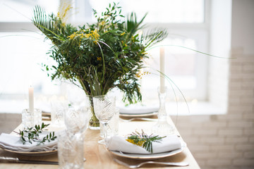 Beautiful springtime table setting with green leaves and mimosa branches, bright white table dinner decoration