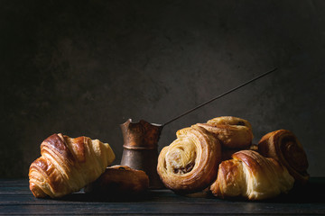 Variety of homemade puff pastry buns cinnamon rolls and croissant served with vintage coffee pot on wooden table. Dark still life. Copy space