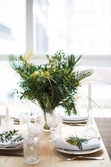 Beautiful springtime table setting with green leaves and mimosa branches, bright white table dinner decoration