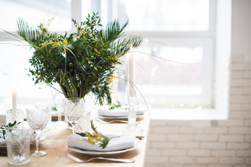 Beautiful springtime table setting with green leaves and mimosa branches, bright white table dinner decoration