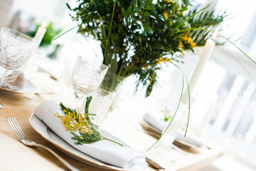 Beautiful springtime table setting with green leaves and mimosa branches, bright white table dinner decoration