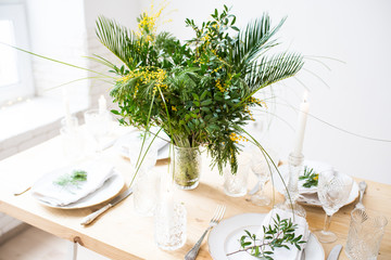 Beautiful springtime table setting with green leaves and mimosa branches, bright white table dinner decoration
