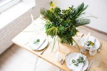 Beautiful springtime table setting with green leaves and mimosa branches, bright white table dinner decoration